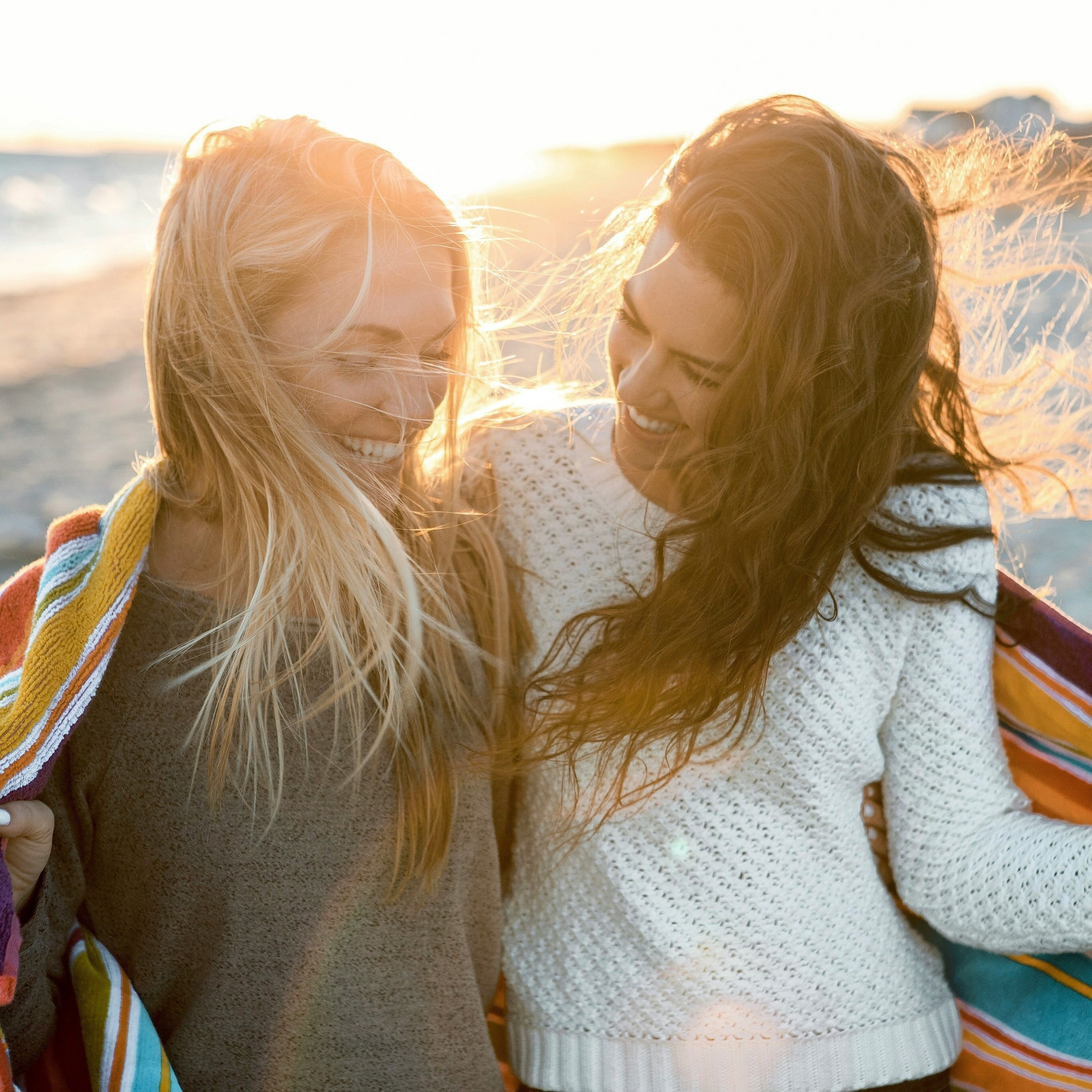 Two women laughing together on a beach at sunset, wrapped in towels.