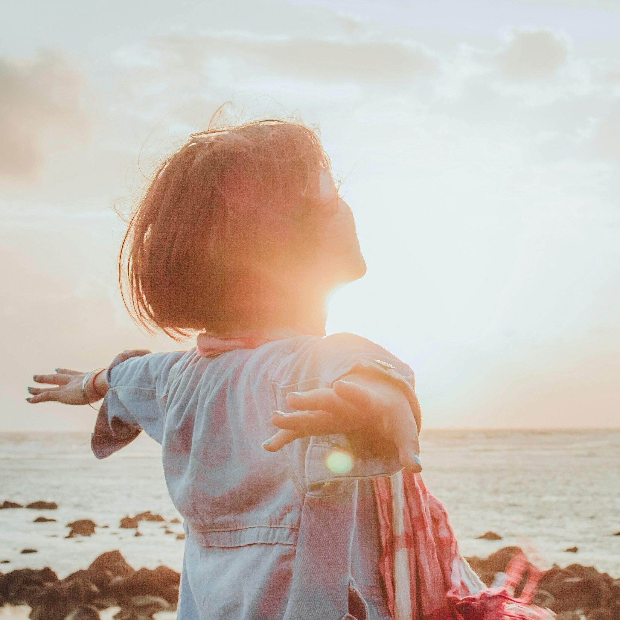Person with short hair standing by the sea, arms outstretched towards a sunset.
